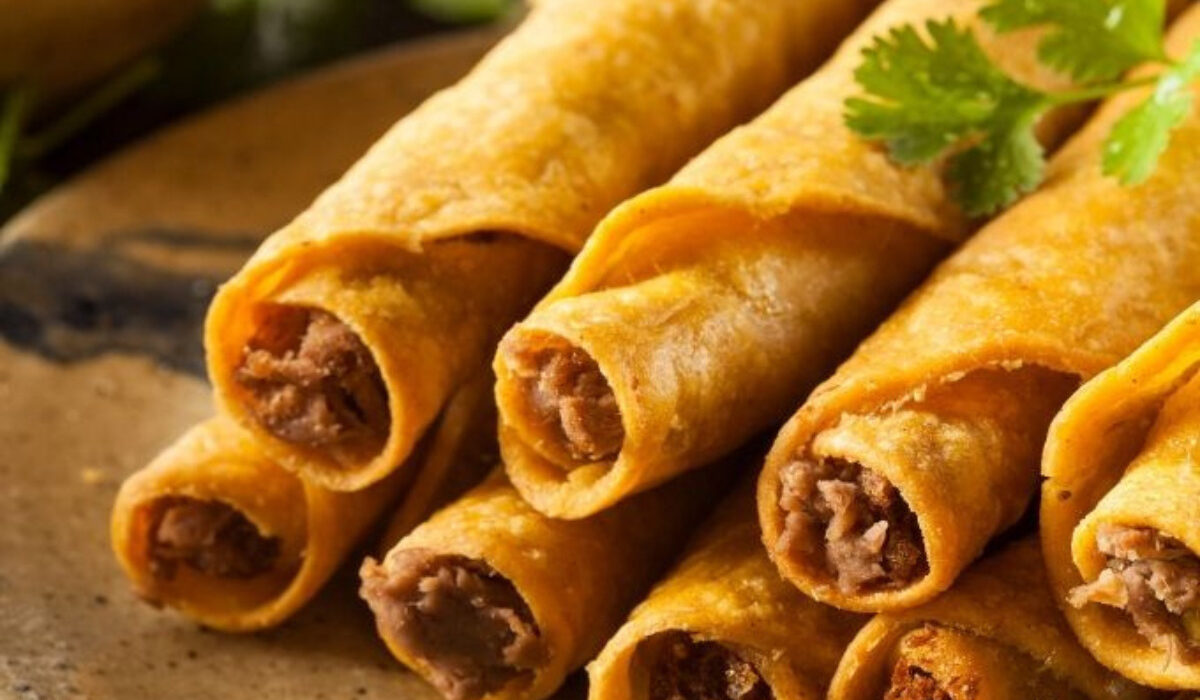 CLose up image of air fryer taquitos stacked on a plate with cilantro and salsa in the background