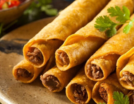 CLose up image of air fryer taquitos stacked on a plate with cilantro and salsa in the background