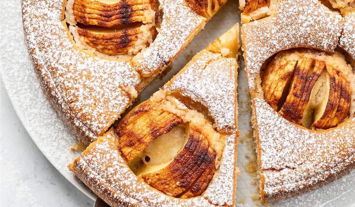 Whole vegan German apple cake dusted with icing sugar and topped with baked apples, with one slice removed and served on a white surface.