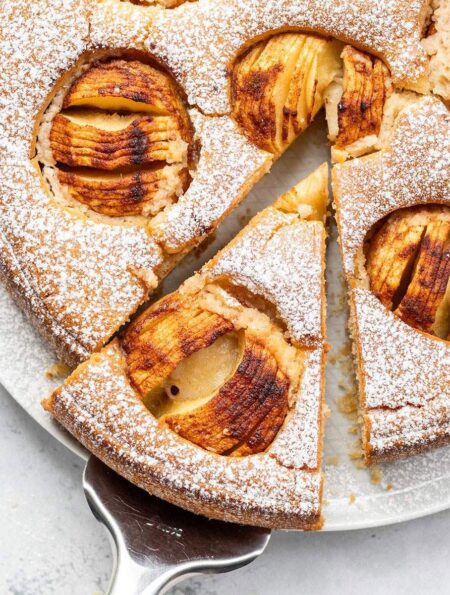 Whole vegan German apple cake dusted with icing sugar and topped with baked apples, with one slice removed and served on a white surface.