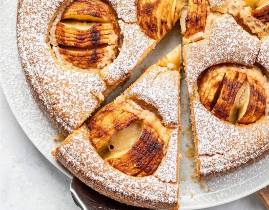 Whole vegan German apple cake dusted with icing sugar and topped with baked apples, with one slice removed and served on a white surface.
