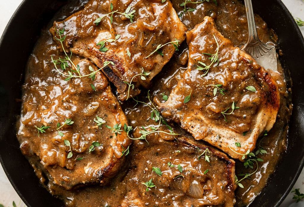 Overhead view of a cast iron pan filled with The Best Smothered Pork Chops topped with fresh herbs and brown gravy.