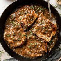 Overhead view of a cast iron pan filled with The Best Smothered Pork Chops topped with fresh herbs and brown gravy.