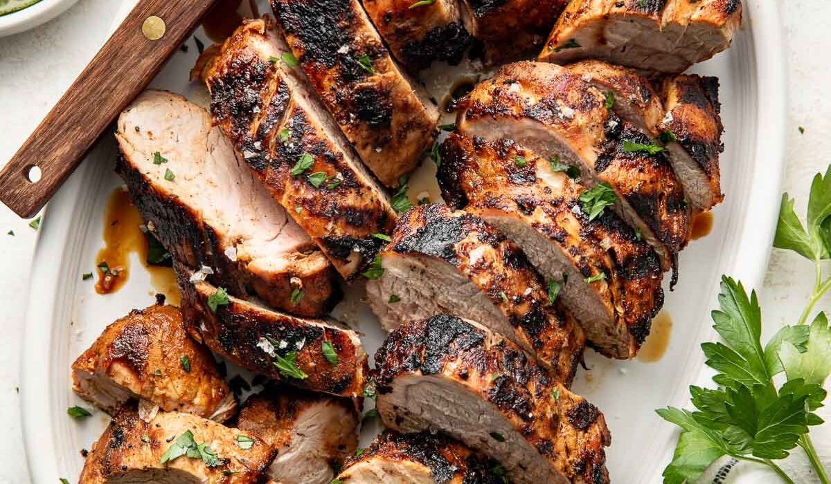 Perfectly grilled and sliced pork tenderloins arranged on a white oval platter surrounded by fresh parsley, limes and red spice mixture in a white bowl.