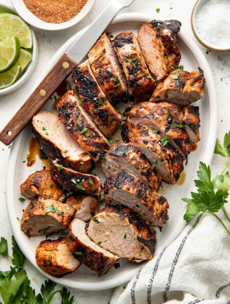 Perfectly grilled and sliced pork tenderloins arranged on a white oval platter surrounded by fresh parsley, limes and red spice mixture in a white bowl.