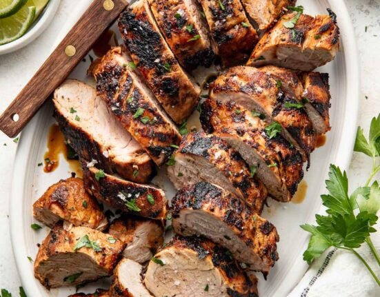 Perfectly grilled and sliced pork tenderloins arranged on a white oval platter surrounded by fresh parsley, limes and red spice mixture in a white bowl.