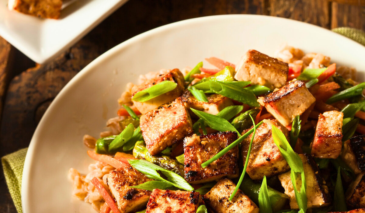 Tofu stir fry on a plate over rice with a serving dish of it next to the plate