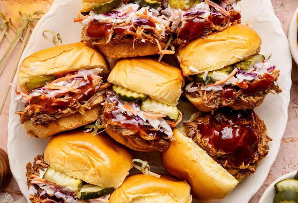 Overhead view of a platter of Pulled Pork Sliders topped with coleslaw and sliced pickles.