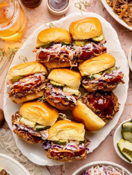 Overhead view of a platter of Pulled Pork Sliders topped with coleslaw and sliced pickles.