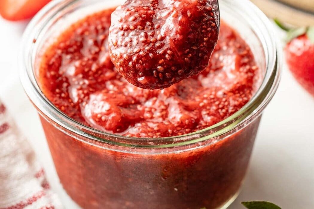 Close up view of a small glass jar of red Chia Seed Jam with a spoon bringing up a scoop.