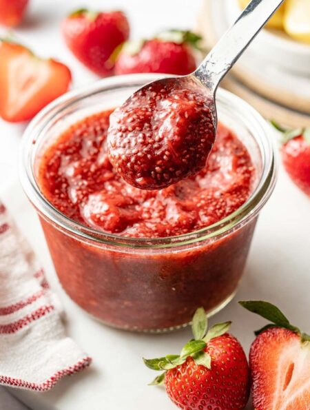 Close up view of a small glass jar of red Chia Seed Jam with a spoon bringing up a scoop.