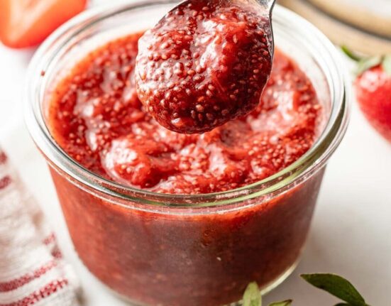Close up view of a small glass jar of red Chia Seed Jam with a spoon bringing up a scoop.