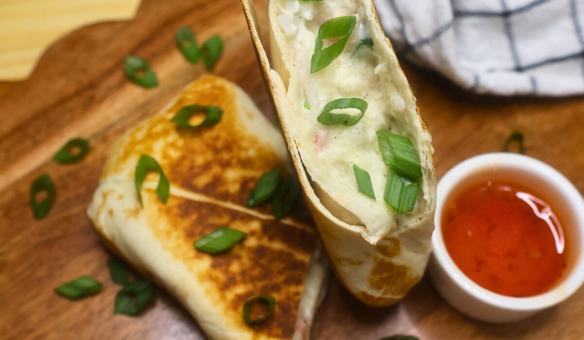 Overhead image of a crab rangoon burrito cut in half on a wooden serving tray with dipping sauce