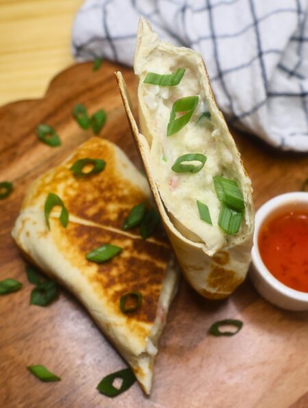 Overhead image of a crab rangoon burrito cut in half on a wooden serving tray with dipping sauce